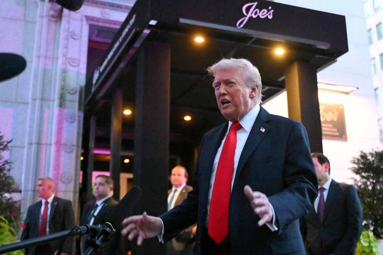 A imagem mostra um homem em pé em frente a um restaurante chamado Joe's. Ele está vestido com um terno escuro e uma gravata vermelha, gesticulando enquanto fala. Ao fundo, há várias pessoas, algumas em trajes formais, e a entrada do restaurante é visível, com iluminação suave. O ambiente parece ser ao ar livre, com plantas ao redor e uma atmosfera de evento.