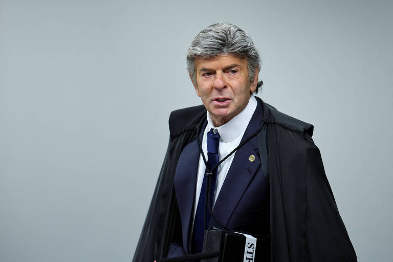 Um homem com cabelo grisalho e bem penteado, vestido com um terno escuro e uma capa preta, está em pé em um ambiente neutro. Ele parece estar se preparando para uma audiência, segurando um objeto que parece ser um livro ou um bloco de notas. O homem tem uma expressão séria e está olhando para a frente.