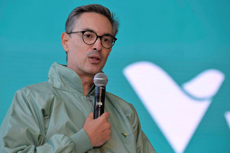 Um homem com cabelo curto e grisalho, usando óculos e uma jaqueta verde, está falando ao microfone em um evento. Ao fundo, há um painel com uma cor de fundo verde e um logotipo estilizado em branco.