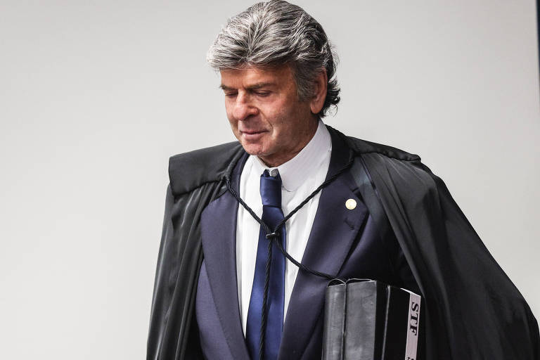 Um homem com cabelo grisalho e liso, usando um terno escuro e uma gravata azul. Ele está vestido com uma capa preta e carrega uma pasta preta. O homem parece estar saindo de um ambiente formal, com uma expressão séria no rosto.
