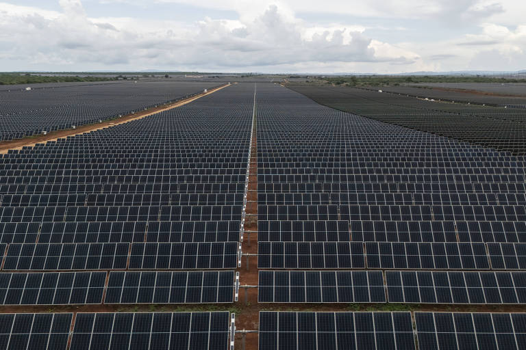 A imagem mostra um vasto campo de painéis solares dispostos em fileiras organizadas, cobrindo uma grande área de terra. O céu está parcialmente nublado, e a paisagem ao fundo é composta por uma extensão de terreno plano.