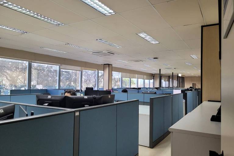 A imagem mostra um ambiente de escritório moderno, com várias estações de trabalho organizadas em cubículos. As paredes dos cubículos são de cor azul clara, e há computadores em cada estação. O espaço é bem iluminado, com janelas grandes que permitem a entrada de luz natural. No fundo, é possível ver mais cubículos e uma área de trabalho vazia.
