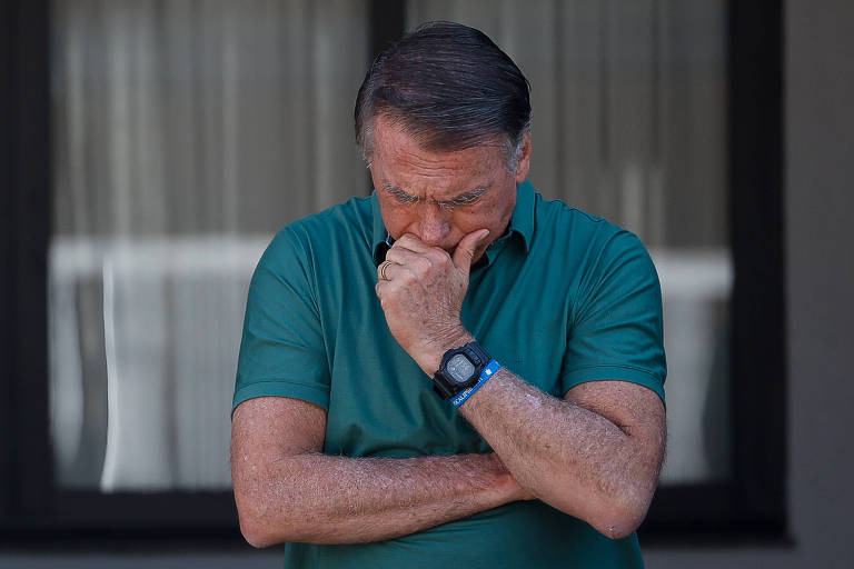 Um homem está em pé, com os braços cruzados e a mão cobrindo a boca, aparentando estar pensativo ou preocupado. Ele usa uma camiseta verde e um relógio no pulso. Ao fundo, há uma janela com cortinas.