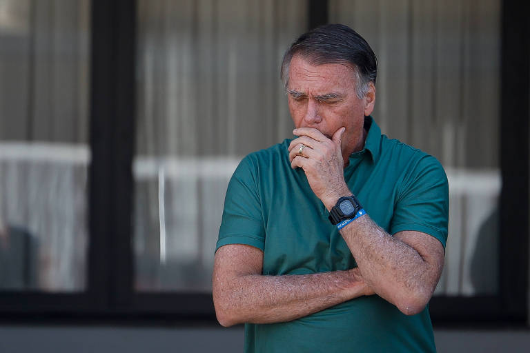 Um homem está em pé, com os braços cruzados e a mão na boca, aparentando estar pensativo. Ele usa uma camisa verde e um relógio no pulso. Ao fundo, há janelas com cortinas que filtram a luz.