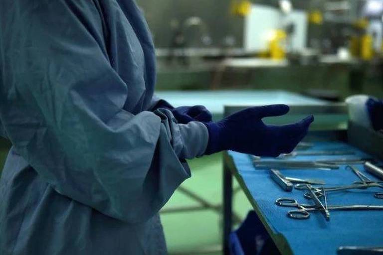Na zdjęciu widać osobę w niebieskim fartuchu chirurgicznym i niebieskich rękawiczkach, przygotowującą się do zabiegu. Ręka osoby jest wyciągnięta, a w tle widać stół z narzędziami chirurgicznymi ułożonymi na niebieskim obrusie. Sceneria wydaje się być salą operacyjną lub centrum chirurgicznym, z odpowiednim oświetleniem i widocznym sprzętem.