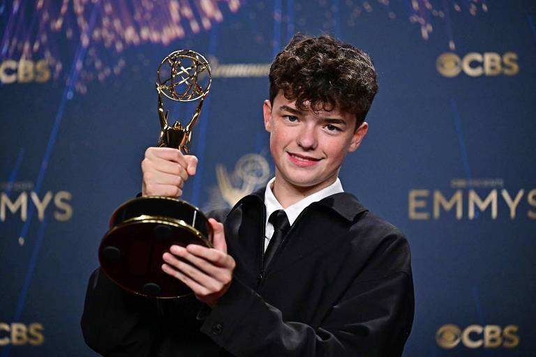 Um jovem está posando para a foto segurando um prêmio Emmy. Ele tem cabelo cacheado e usa uma camisa branca com um paletó preto. O fundo é decorado com elementos que lembram uma cerimônia de premiação, com o logotipo da CBS e a palavra 'EMMYS'.
