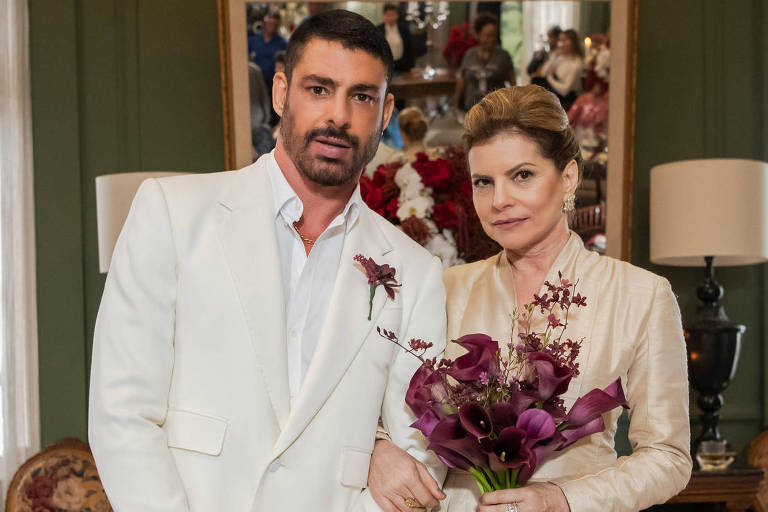 Um homem e uma mulher posam juntos em um ambiente elegante. O homem está vestido com um terno branco e tem cabelo escuro e barba. A mulher usa um vestido claro e segura um buquê de flores roxas. Ao fundo, há uma parede decorada com quadros e uma iluminação suave, além de móveis clássicos como cadeiras e mesas.