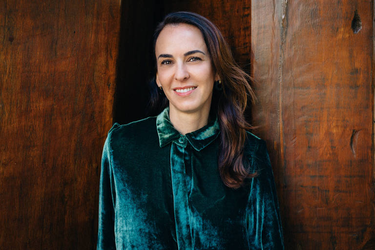 A imagem mostra uma mulher sorrindo, com cabelo solto e levemente ondulado. Ela está vestindo uma blusa de veludo verde com um colarinho. O fundo é composto por painéis de madeira escura, criando um contraste com a roupa da mulher. A iluminação é suave, destacando seu rosto e a textura do tecido.