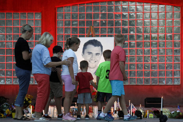 Um grupo de adultos e crianças se reúne em frente a um retrato emoldurado de um homem, que está exposto em um local com paredes de vidro. As pessoas estão em um ambiente de homenagem, com velas acesas e flores ao redor. As crianças estão em destaque, observando o retrato, enquanto os adultos parecem estar confortando umas às outras. O cenário é iluminado de forma suave, criando uma atmosfera de respeito e lembrança.