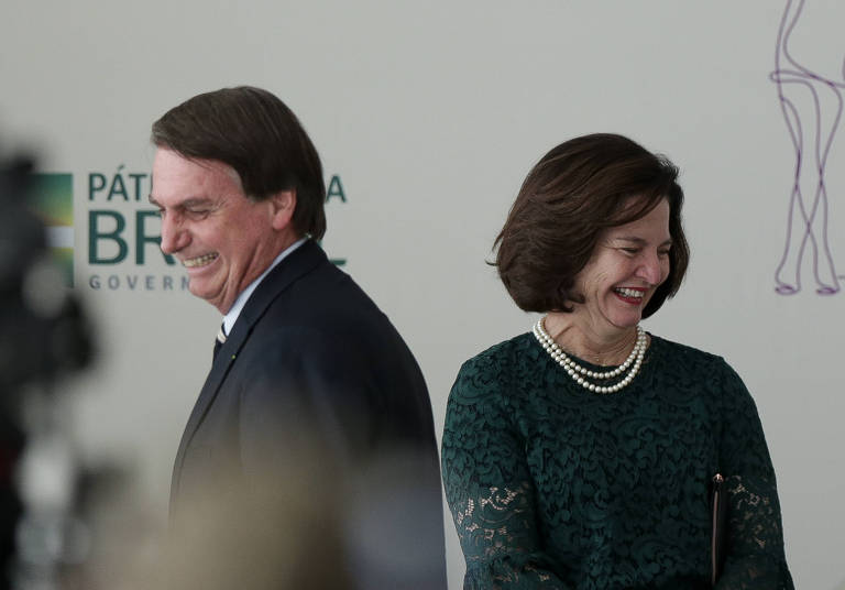 Um homem e uma mulher, sorridentes, ambos vestindo roupas escuras, de costas, um para o outro, distribuem cumprimentos no palco de uma solenidade, Atrás, um cartaz do evento.