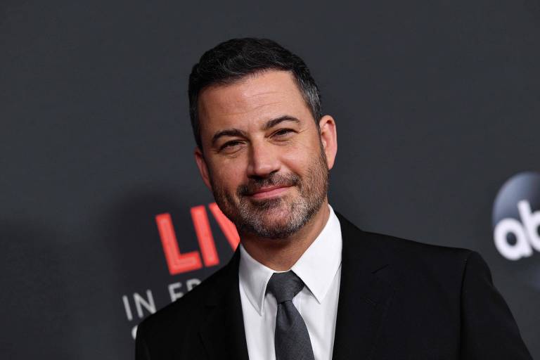 Um homem com cabelo escuro e barba, vestindo um terno preto e gravata, está posando para a câmera em um evento. O fundo é escuro com texto em vermelho que diz 'LIVE IN FRONT OF A STUDIO AUDIENCE'.