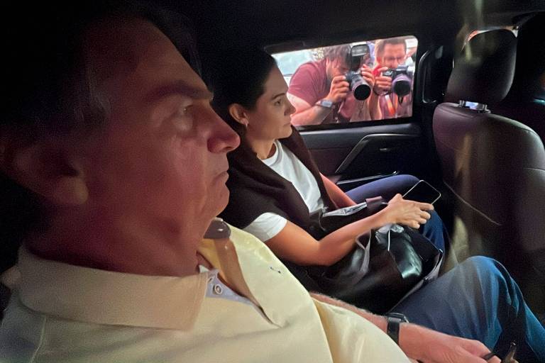 A imagem mostra duas pessoas dentro de um veículo. À esquerda, um homem com cabelo curto e uma camisa polo clara, aparentando estar pensativo. À direita, uma mulher com cabelo preso, usando uma blusa escura e segurando um celular. Ao fundo, é possível ver fotógrafos tirando fotos através da janela do carro.