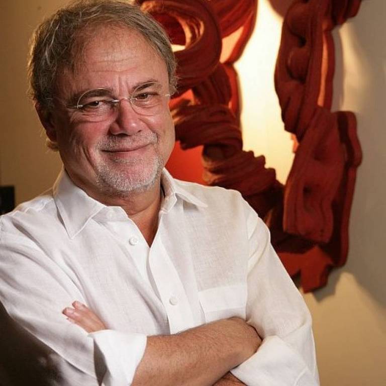 Um homem de cabelos grisalhos e barba, usando uma camisa branca, está posando com os braços cruzados. Ele sorri levemente e está em frente a uma parede decorada com uma obra de arte abstrata em tons de vermelho. O ambiente parece ser bem iluminado.