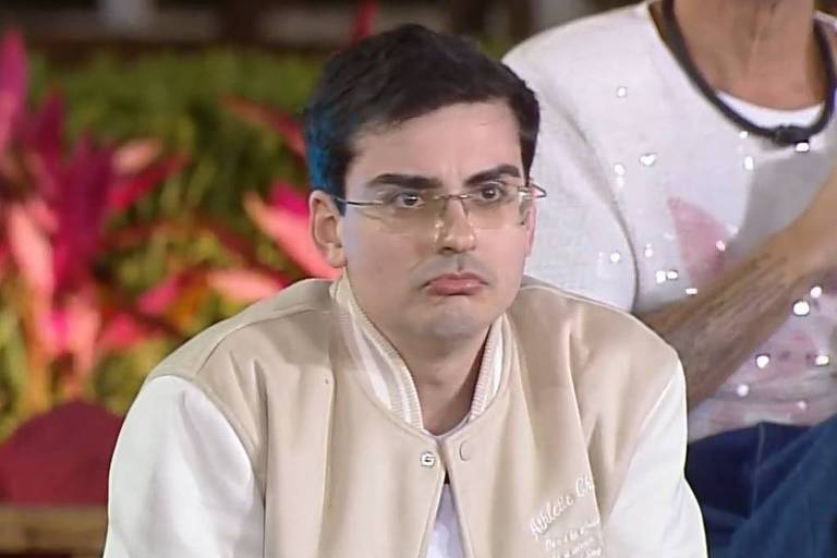 Um homem com óculos e cabelo escuro, usando uma jaqueta clara com detalhes em branco, está sentado em um ambiente com plantas coloridas ao fundo. Ele parece pensativo ou preocupado, com uma expressão facial neutra. Ao fundo, há outras pessoas, mas não estão claramente visíveis.