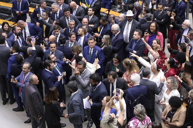 A imagem mostra um grande grupo de pessoas reunidas em um ambiente que parece ser o Congresso Nacional do Brasil. Há muitos homens e mulheres, a maioria vestindo ternos e roupas formais, interagindo entre si. Algumas pessoas estão segurando celulares, tirando fotos ou gravando. O ambiente é bem iluminado e há uma sensação de agitação e atividade. Ao fundo, é possível ver uma escadaria e algumas bandeiras do Brasil.