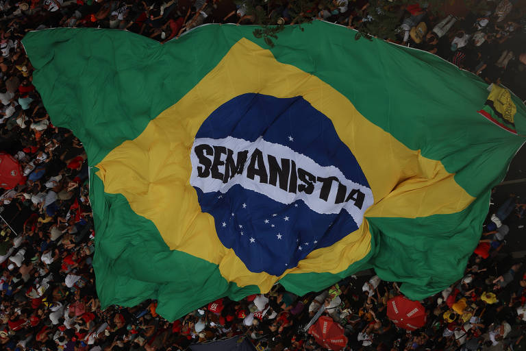 A imagem mostra uma grande bandeira do Brasil sendo exibida em um evento, com o fundo repleto de pessoas. A bandeira é predominantemente verde e amarelo, com um grande losango azul no centro, contendo estrelas brancas e a inscrição 'Ordem e Progresso' em uma faixa branca. A cena transmite um sentimento de patriotismo e celebração.