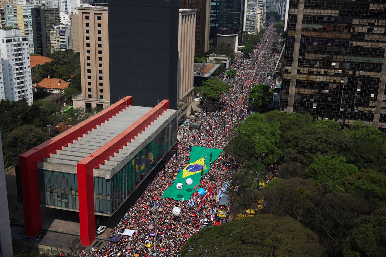 A imagem mostra uma grande manifestação em uma avenida de São Paulo, com uma multidão de pessoas reunidas. No centro da imagem, há um grande painel verde e amarelo, simbolizando a bandeira do Brasil. Ao fundo, é possível ver prédios altos e árvores ao longo da avenida. A cena é marcada por um clima de agitação e união entre os manifestantes.