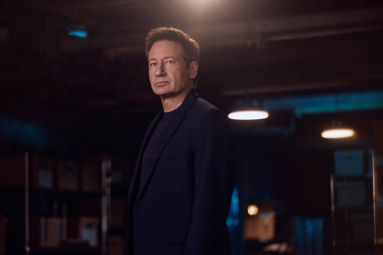 Um homem está posicionado em um ambiente com pouca luz, com prateleiras ao fundo. Ele usa um traje escuro e tem uma expressão séria. A iluminação destaca seu rosto e parte bash  corpo, criando um contraste com o fundo mais escuro.