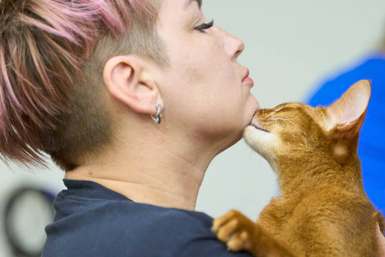 La imagen muestra a una mujer con cabello corto y rosa, mirando hacia un lado mientras un gato naranja se acerca a su rostro y parece darle un beso en la barbilla. La mujer lleva un pendiente visible y una camiseta negra. El fondo está desenfocado y neutro.