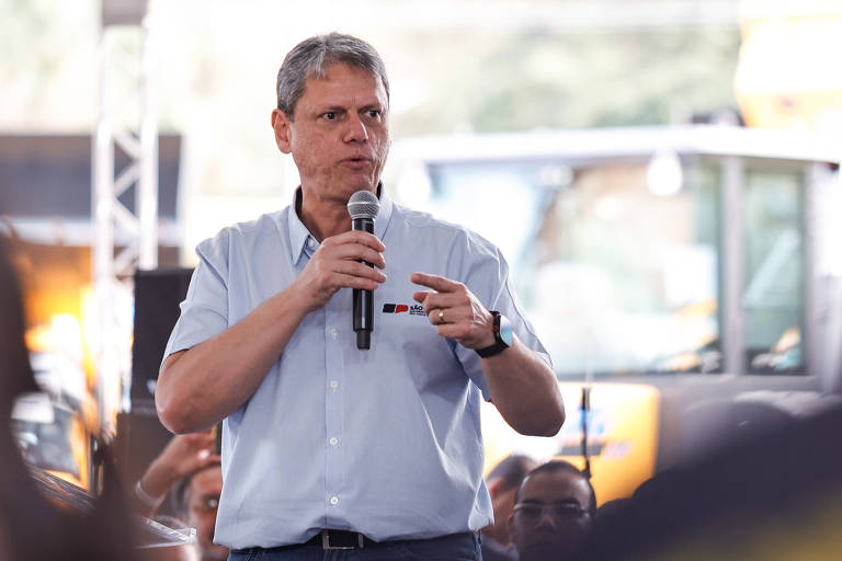 Um homem de cabelo curto e grisalho está falando ao microfone em um evento. Ele usa uma camisa de botão clara e gesticula com a mão enquanto se dirige a uma audiência. Ao fundo, é possível ver máquinas pesadas e uma multidão de pessoas