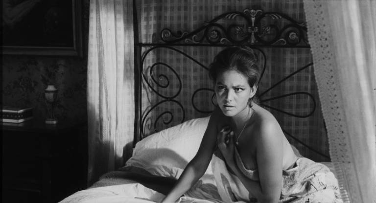 A imagem em preto e branco mostra uma mulher sentada em uma cama, com um olhar pensativo e sério. Ela está vestindo uma blusa de alças e parece estar em um ambiente interno, com paredes decoradas e cortinas. O fundo inclui uma cabeceira de cama de ferro e uma mesa com objetos, sugerindo um ambiente doméstico.