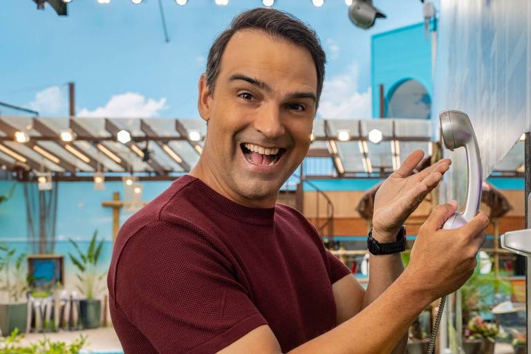 Um homem sorridente está em um ambiente externo, próximo a uma parede decorada. Ele está segurando um telefone de parede e parece estar se divertindo. O fundo apresenta uma área verde com plantas e uma piscina visível ao fundo. O homem usa uma camiseta de cor escura e tem cabelo curto e liso.