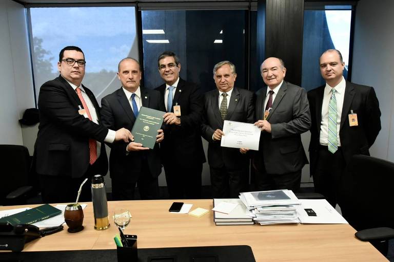 A imagem mostra um grupo de cinco homens em um ambiente de escritório. Eles estão em torno de uma mesa, onde há documentos e uma bebida. Um dos homens, que está no centro, segura um documento verde, enquanto outro homem à sua direita segura um papel. Todos estão vestidos formalmente e parecem estar em uma reunião. Ao fundo, há janelas que mostram um dia claro.