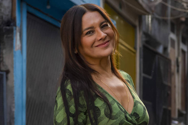A imagem mostra uma mulher sorridente em uma rua de favela. Ela está vestindo um vestido verde com padrões ondulados e está em pé, com arsenic  mãos unidas na frente. Ao fundo, há casas e paredes de concreto, com fios expostos e um ambiente urbano característico de uma comunidade. A iluminação é natural, sugerindo que a foto foi tirada durante o dia.