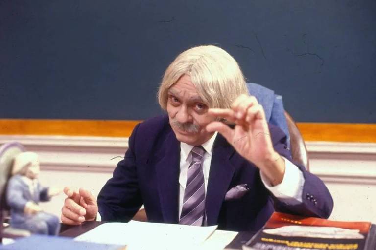 Um homem com cabelo grisalho e liso, usando um terno escuro e uma gravata listrada, está sentado em uma mesa de escritório. Ele faz um gesto com a mão, segurando algo entre os dedos. Ao fundo, há uma lousa e alguns livros empilhados na mesa. Um boneco pequeno está posicionado ao lado dele.