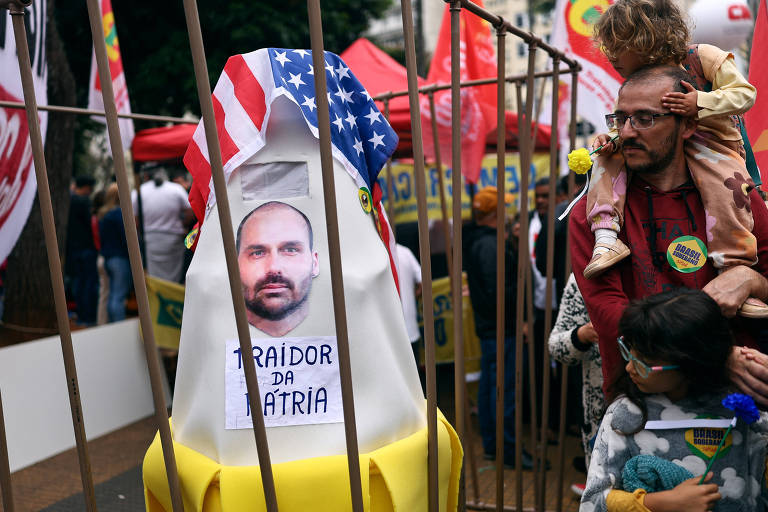 A imagem mostra um protesto onde uma figura central  um boneco com uma foto de um homem e a inscrio "TRAIDOR DA PTRIA". O boneco est cercado por uma grade, e ao fundo h pessoas participando do protesto, algumas segurando bandeiras e cartazes. Uma criana est nas costas de um dos manifestantes, que parece estar segurando uma flor. O ambiente  de agitao, com tendas e bandeiras visveis.