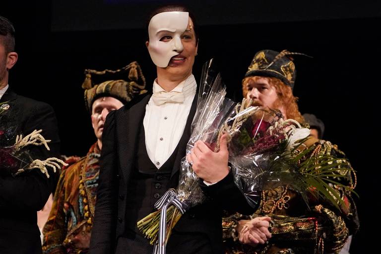 Um grupo de atores em um palco, com um foco principal em um homem usando uma máscara de fantasma. Ele está vestido com um traje formal e segura um buquê de flores. Ao fundo, outros atores estão vestidos com trajes elaborados, incluindo um com um chapéu decorado. A cena parece ser de uma apresentação teatral, possivelmente de 'O Fantasma da Ópera'.