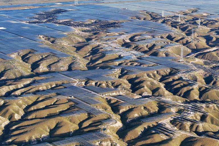 A imagem mostra uma vasta área de painéis solares dispostos em um terreno montanhoso. As ondulações do solo são visíveis entre os painéis, que refletem a luz do sol. Linhas de cabos e torres de transmissão também podem ser vistas ao fundo, indicando a infraestrutura elétrica associada à geração de energia solar.