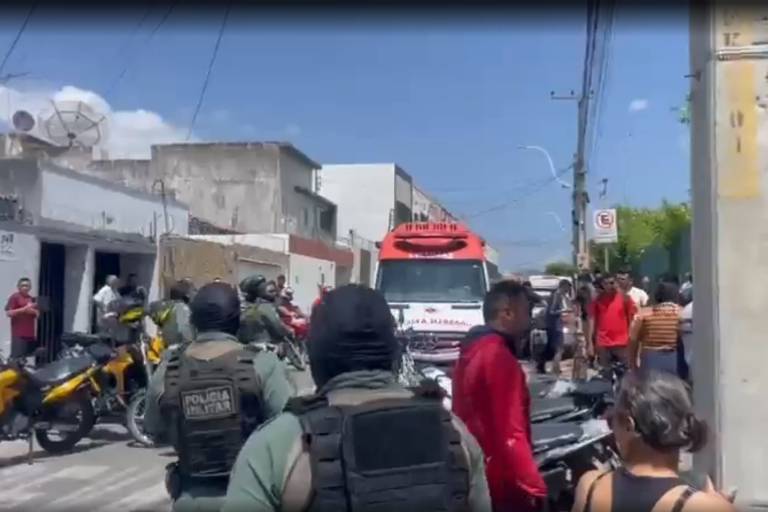 A imagem mostra uma cena de intervenção policial em uma rua urbana. Há vários policiais armados e com coletes à prova de balas, alguns deles de costas para a câmera. Um veículo de emergência, possivelmente uma ambulância, está estacionado na rua. Há uma multidão de pessoas observando a cena, algumas em pé e outras sentadas em motocicletas. O céu está claro e ensolarado, e as construções ao fundo são de um estilo simples, com paredes brancas e algumas janelas.