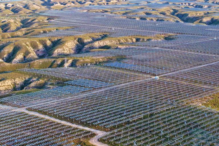 A imagem mostra uma vasta área coberta por painéis solares dispostos em linhas organizadas, com colinas ao fundo. O terreno é irregular, com ondulações visíveis, e os painéis refletem a luz do sol, criando um padrão geométrico na paisagem. A vegetação é escassa, predominando a terra exposta entre os painéis.