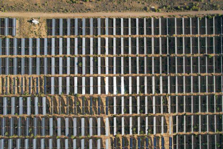 A imagem mostra uma grande área coberta por painéis solares dispostos em linhas retas. Os painéis são predominantemente de cor escura, com algumas seções mais claras. O terreno ao redor é árido, com vegetação escassa visível entre os painéis. No canto superior esquerdo, há uma estrutura pequena, possivelmente um veículo ou equipamento relacionado à instalação.