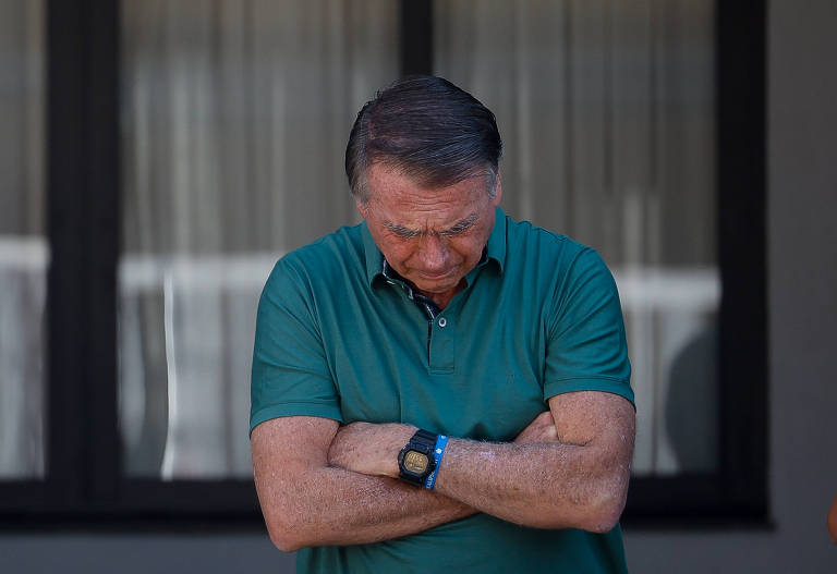 Un hombre está de pie con los brazos cruzados y la cabeza gacha, expresando reflexión o tristeza. Lleva un polo verde y un reloj en la muñeca. Al fondo se ve una ventana con cortinas.
