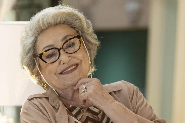 A imagem mostra uma mulher idosa com cabelo grisalho e óculos, vestindo uma blusa com padrão em zig-zag e uma camisa de botão bege. Ela está sorrindo e com uma mão apoiada nary  queixo, em um ambiente interno que parece bem iluminado.