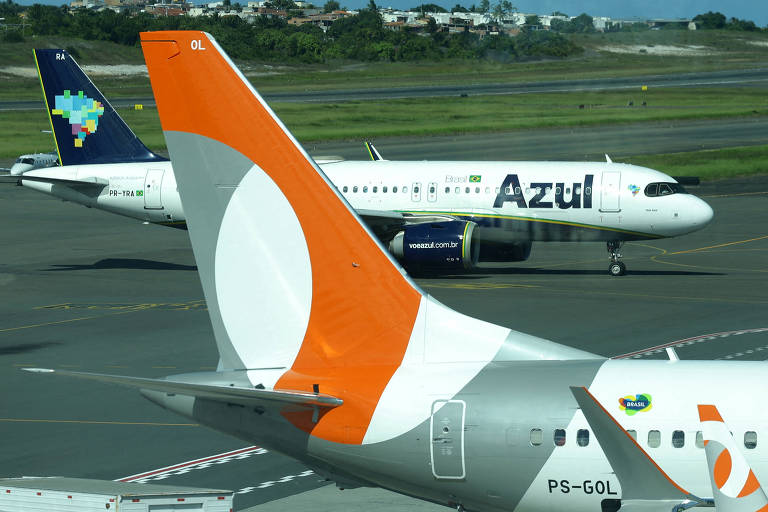 La cola de un avión Gol, naranja y blanco, en primer plano. Al fondo, un avión Azul con el logo azul y verde rodando por la pista.