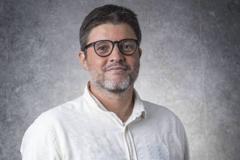 Man with abbreviated  acheronian  hair, beard, and glasses wearing a achromatic  button-up garment  stands against a grey  textured background.