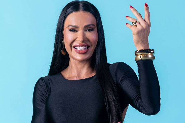 A imagem mostra uma mulher sorridente vestindo um vestido preto justo. Ela tem cabelo longo e liso, e está usando brincos e várias pulseiras douradas. A mulher está posando com uma das mãos levantadas, em um fundo azul claro.