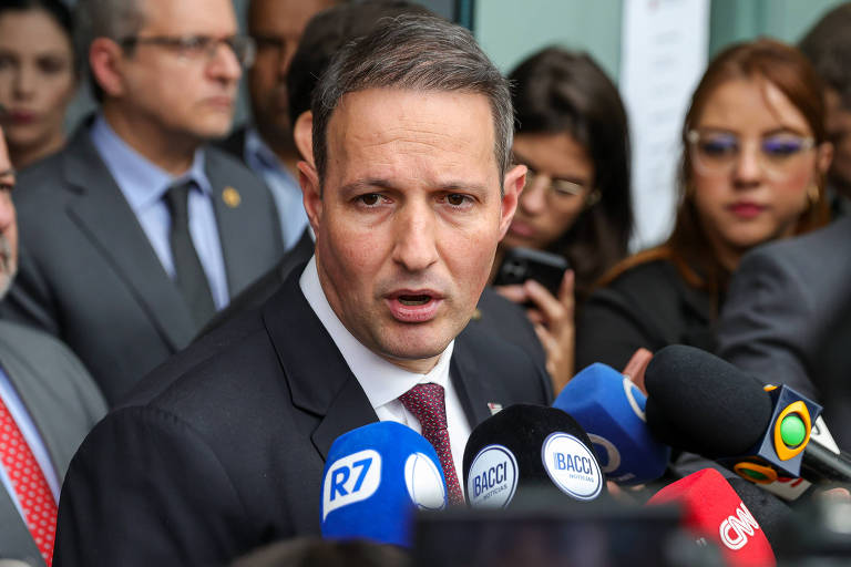 Um homem em um terno escuro está falando em uma coletiva de imprensa, cercado por jornalistas e câmeras. Ele parece estar respondendo a perguntas, com microfones de várias emissoras de televisão à sua frente. Ao fundo, há outras pessoas ouvindo atentamente, algumas com expressões sérias. O ambiente parece ser um local público, possivelmente fora de um edifício governamental.