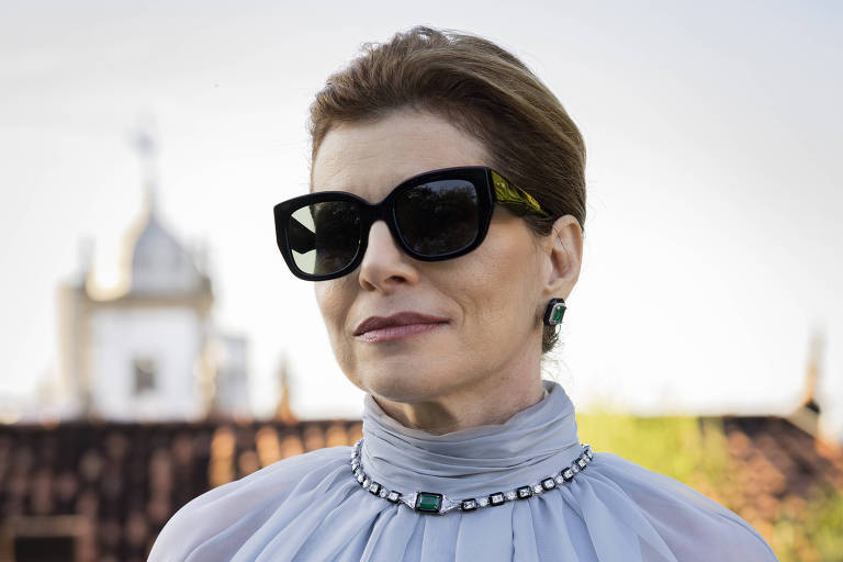 Uma mulher está usando um vestido longo e elegante de cor cinza claro, com mangas longas e um colar de corrente ao redor bash  pescoço. Ela está usando óculos escuros e tem o cabelo preso. Ao fundo, há telhados e uma construção com cúpula, sugerindo um ambiente urbano.