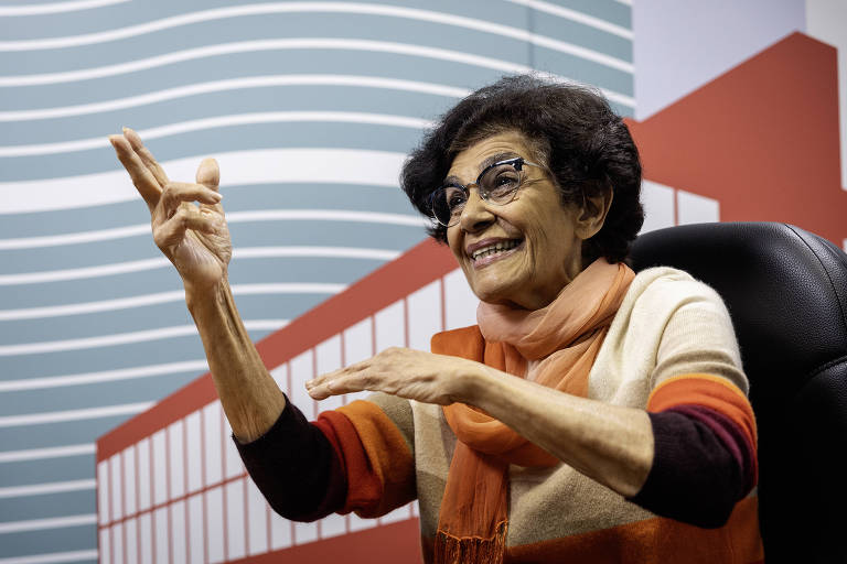 Mulher idosa com cabelo curto e óculos, vestindo suéter listrado em tons de bege, laranja e marrom, e cachecol laranja, sentada em cadeira preta, gesticulando com as mãos. Fundo apresenta padrão gráfico com linhas curvas brancas sobre azul e formas geométricas vermelhas e brancas.