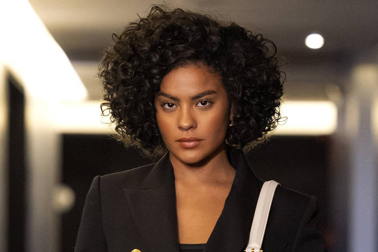 Uma mulher com cabelo cacheado e volumoso está caminhando por um corredor. Ela usa um blazer preto com botões dourados e calças pretas justas. A mulher carrega uma bolsa branca e parece confiante. O corredor tem paredes escuras e iluminação suave, com portas em ambos os lados.