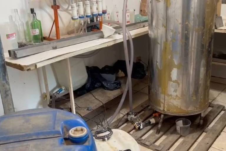 Tanque metálico cilíndrico com sinais de desgaste apoiado sobre paletes de madeira em chão de azulejos. Ao lado, bancada branca com garrafas, frascos e tubos conectados ao tanque. Ambiente interno com paredes claras e objetos diversos ao fundo.