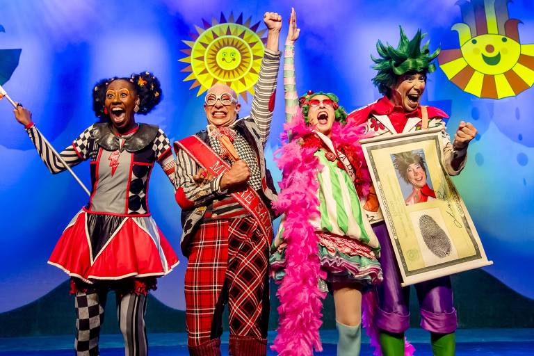 Four actors in vibrant costumes and excited expressions line up on stage. From left to right: Woman wearing red and black dress, striped stockings, hair in two buns; A man wearing glasses, a red headband, and plaid pants; A woman with a pink wig, pink feathers, and a colorful dress; And a man with a spiky green wig and a red shirt, holding a frame with a photo and a fingerprint on it. The blue background has smiling sun designs.