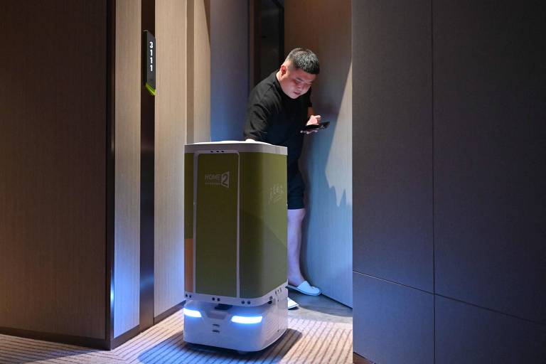 Robô de entrega autônomo com luzes azuis na base circula corredor de hotel enquanto hóspede em roupa casual preta usa celular próximo à porta de quarto.