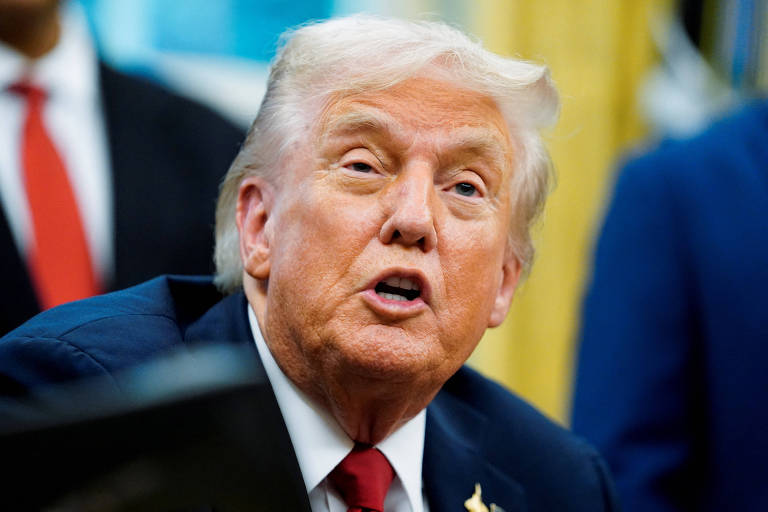 Donald Trump, ex-presidente dos Estados Unidos, aparece em close, vestindo terno azul escuro, camisa branca e gravata vermelha. Fundo desfocado com pessoas e decoração típica de escritório oficial.