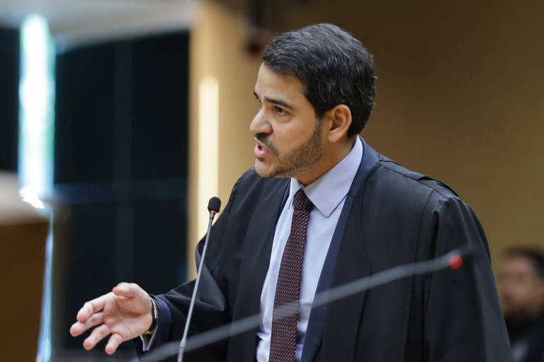 Advogado de terno, gravata e toga preta fala com expressão séria em tribunal, gesticulando com a mão direita próximo a microfone.