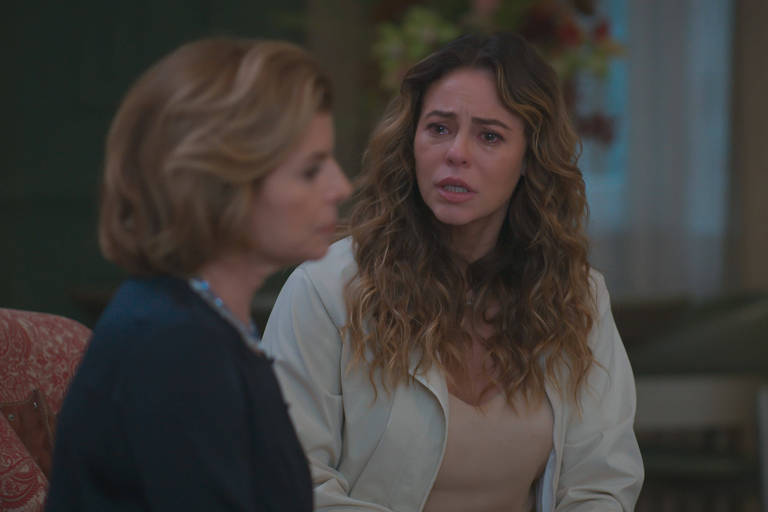 Duas mulheres sentadas em um ambiente interno. A mulher à direita, com cabelos longos e ondulados, veste casaco claro e expressão séria. A mulher à esquerda, com cabelo curto e loiro, está desfocada e veste roupa escura.